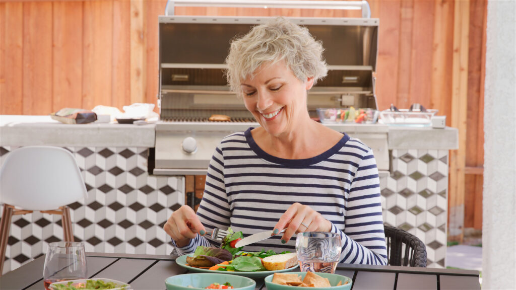 Senior Woman Outdoors At Home Sitting At Table Eating Barbecue Meal With Salad